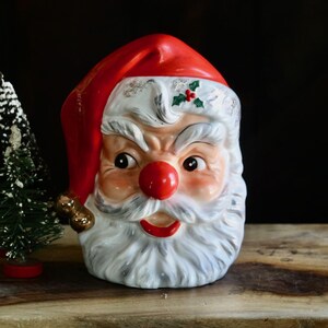 May include: A vintage Santa Claus head figurine with a red hat and rosy cheeks. The figurine has a white beard and mustache, and a red nose. A small Christmas tree is to the left of the Santa head. The items are on a wooden surface.
