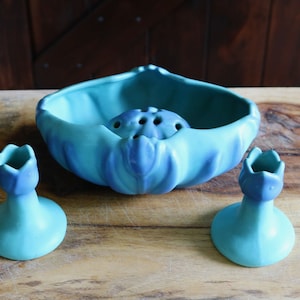 May include: A vintage ceramic flower frog set in shades of turquoise and blue. The set includes a large bowl with a built-in flower holder and two small candle holders. The pieces are arranged on a wooden surface.
