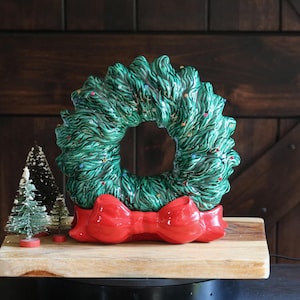May include: A ceramic Christmas wreath featuring textured green pine needles with small multi-colored accents and a large, smooth, glossy red bow at the base. Next to the wreath are three small, conical, bottle-brush style decorative trees.