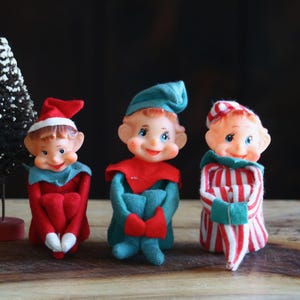 May include: Three vintage Christmas elf figurines with smiling faces, wearing red, green, and striped outfits. Two small Christmas trees are in the background, one with snow. The elves are posed on a wooden surface.