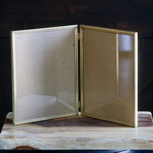 May include: A gold-colored, hinged picture frame, open to display two photos. The frame has a simple, rectangular design with a thin border. The background is a dark wooden surface.