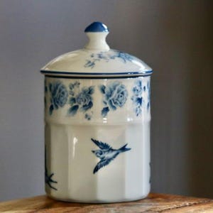 May include: A white ceramic jar with a lid, adorned with blue floral patterns and a flying bird design. The jar has a cylindrical shape with a lid and a small blue knob. The floral pattern includes roses. The jar is sitting on a wooden surface.