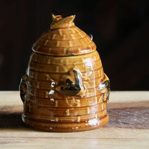 May include: A ceramic honey pot with a lid, shaped like a beehive. The pot is a warm brown color with darker brown accents and a glossy finish. The beehive design includes textured horizontal lines and bee-shaped details.