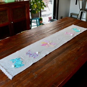 May include: A beige Easter table runner with appliqued bunnies and a flower design. The runner has a scalloped edge and is decorated with small white pom-poms. The runner is placed on a dark brown wooden table.