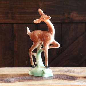 May include: A ceramic deer figurine in shades of brown and white, standing on a light green base with a stylized plant. The deer has a slender neck and is posed with one leg raised. The figurine is likely vintage.