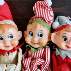 May include: Three vintage elf dolls with smiling faces and rosy cheeks. The elves wear red and green outfits, including pointed hats with white pom-poms. One elf has a red and white striped hat and outfit.