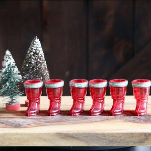 May include: Six red, boot-shaped shot glasses with a green and white trim are arranged on a wooden board. Miniature Christmas trees are in the background. The shot glasses are a festive holiday decoration.
