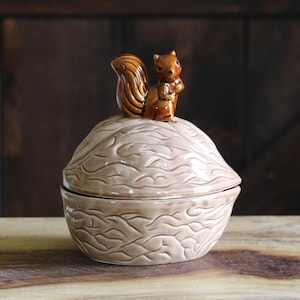 May include: A ceramic nut-shaped container with a brown squirrel figurine on top. The container is light brown with a textured surface, and the squirrel is brown with a bushy tail. The container is likely used for storing small items.