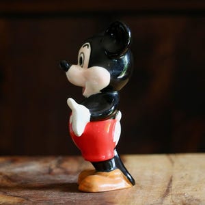 May include: A vintage Mickey Mouse figurine. The ceramic figure features a black head and ears, a white face, hands, and gloves, red shorts, and tan shoes. The figurine is posed in a classic stance, with one hand raised.