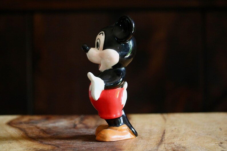 May include: A vintage Mickey Mouse figurine. The ceramic figure features a black head and ears, a white face, hands, and gloves, red shorts, and tan shoes. The figurine is posed in a classic stance, with one hand raised.