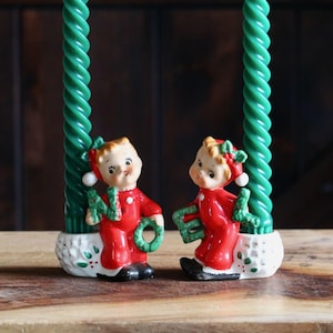 May include: Two vintage Christmas candle holders featuring ceramic figures of children in red outfits, holding green wreaths and the letters "N" and "E". The figures are positioned in front of green, spiral-shaped candles.