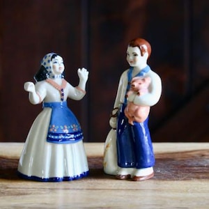 May include: A pair of ceramic figurines depicting a man and woman in traditional attire. The woman wears a white dress with a blue apron and headscarf. The man holds a piglet. The figurines are set against a dark wooden background.