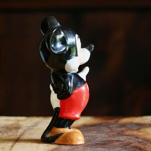 May include: A vintage Mickey Mouse figurine. The figure is primarily black, with white gloves and face, and red shorts. The base is tan. The figurine is posed in a side view, with one hand raised.
