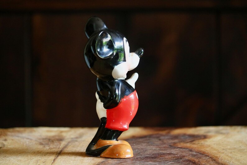 May include: A vintage Mickey Mouse figurine. The figure is primarily black, with white gloves and face, and red shorts. The base is tan. The figurine is posed in a side view, with one hand raised.