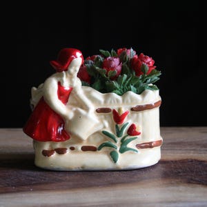 May include: A ceramic planter featuring a woman in a red dress watering flowers. The planter is cream-colored with brown accents and a floral design. The flowers are red and green, adding a pop of color to the piece.