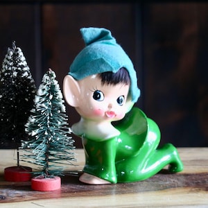 May include: A vintage ceramic elf figurine with a teal hat and green outfit, kneeling on a wooden surface. Two small Christmas trees with snow-covered branches stand nearby, adding a festive touch to the scene.