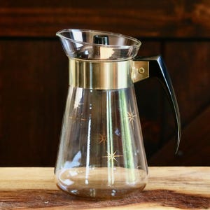 May include: A clear glass carafe featuring a gold-toned metal band around the neck and a black plastic handle. The glass body is decorated with gold starburst patterns. A black plastic lid is fitted into the top opening of the glass vessel.