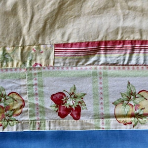 May include: Close-up of a fabric with a patchwork design. The top section is a solid pale yellow. Below, a section with red and white stripes, and a section with a fruit pattern of apples and strawberries on a white and green checkered background.