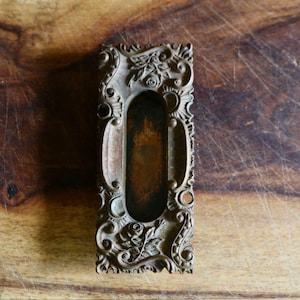 May include: Ornate antique bronze-toned drawer pull with a rectangular shape and a central recessed handle. The pull is decorated with floral and scrollwork designs, showcasing a vintage aesthetic. The hardware is set against a wooden surface.