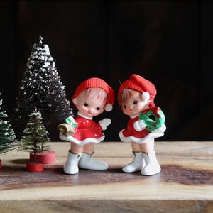 May include: Two ceramic figurines of children wearing red hats, red dresses, and white boots. One child holds a gold bell, and the other holds a green wreath. Next to them are two miniature artificial pine trees with white snow-like tips.