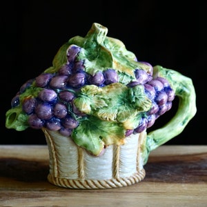 May include: A ceramic teapot shaped like a basket of grapes. The teapot features a white basket with a woven texture, green leaves, and purple grapes. The handle and spout are green.