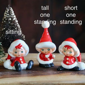 May include: A festive holiday scene featuring three ceramic elf figurines in red and white outfits, with black shoes. Two small Christmas trees are also present. The text on the image reads "tall one standing", "short one standing", and "sitting".