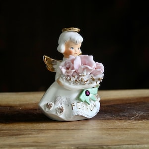 May include: A ceramic angel figurine with white hair, a gold halo, and gold wings. The angel wears a white dress with gold accents and holds a bouquet of pink roses. The word FEBRUARY is printed on the front of the dress, which features a small green bow with a purple gem.