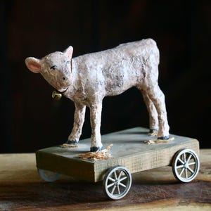 May include: A handcrafted, rustic-style toy featuring a light-colored calf figurine with a bell around its neck, mounted on a wooden cart with four small wheels. The calf has a textured appearance, and the cart has a weathered look.