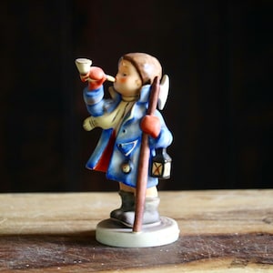 May include: A vintage figurine of a child in a blue coat, red gloves, and a white scarf, blowing a horn. The figurine holds a wooden staff and a lantern. The base is a light green color.