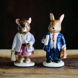 May include: Two ceramic figurines of anthropomorphic animals. One is a mouse wearing a lavender jacket and burgundy skirt, holding a scroll and a bag. The other is a rabbit in a blue suit, holding a book and a briefcase. Both are on white bases.