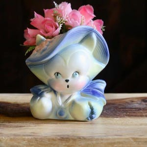 May include: A vintage ceramic cat-shaped planter, painted in pastel blue, yellow, and white, with a blue hat and bow. The cat planter holds pink artificial flowers. The planter is sitting on a wooden surface.
