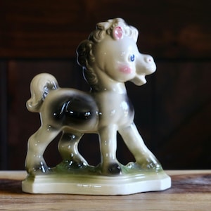 May include: A ceramic horse figurine in a standing pose. The horse has a white body with a black patch on its back, pink cheeks, blue eyes, and a tan mane and tail. It stands on a green base.