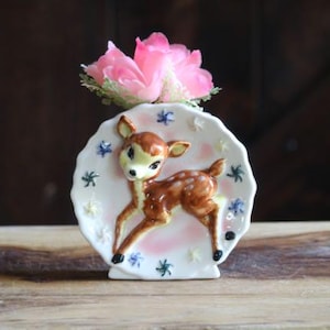 May include: A vintage ceramic vase featuring a brown and white deer with a pink and white background. The vase is decorated with blue and yellow star-shaped accents. A pink flower sits atop the vase.