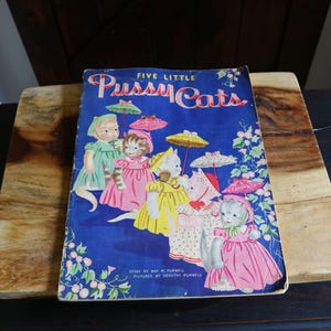 May include: Vintage children's book titled "Five Little Pussy Cats" with a blue cover. The cover features illustrations of five anthropomorphic cats wearing dresses and holding umbrellas. The book is resting on a wooden cutting board.