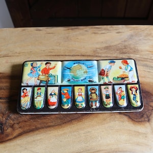 May include: Vintage metal paint box featuring illustrations of children, travel, and world cultures. The box has a rectangular shape with a black border and a wooden background. The box is decorated with colorful images.
