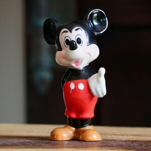 May include: A vintage Mickey Mouse figurine. The figure is depicted in a classic pose, with a black body, red shorts, and tan shoes. The figurine has a white gloved hand raised in a thumbs-up gesture. The background is blurred, with a wooden surface.