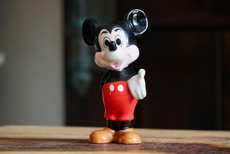 May include: A vintage Mickey Mouse figurine. The figure is depicted in a classic pose, with a black body, red shorts, and tan shoes. The figurine has a white gloved hand raised in a thumbs-up gesture. The background is blurred, with a wooden surface.
