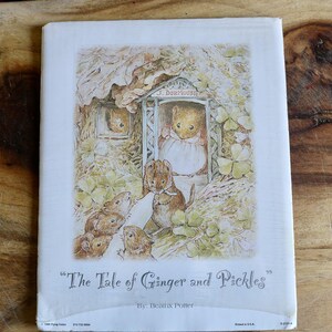 May include: A vintage book cover titled "The Tale of Ginger and Pickles" by Beatrix Potter. The illustration features several mice in a whimsical setting, with a mouse in a doorway and others gathered around. The cover is white with a colorful illustration.
