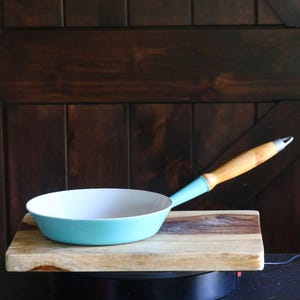 May include: A small, turquoise-colored frying pan with a light wood handle and a white interior. The pan rests on a wooden cutting board. The handle is light wood with a metal end.