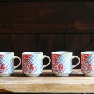 May include: Four ceramic mugs with a patchwork design in red, blue, and green floral patterns. The mugs have white handles and bases, set on a wooden board against a dark wood background.