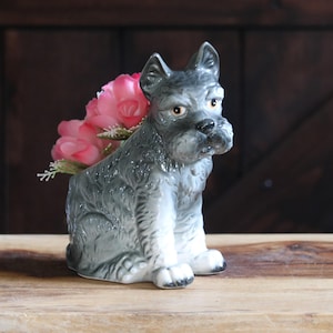 May include: A ceramic figurine of a seated gray and white schnauzer dog with a textured coat and alert expression. The dog figurine features an open top section containing a cluster of artificial pink roses with green stems and foliage.