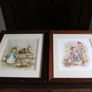May include: Two framed prints featuring Beatrix Potter's Peter Rabbit illustrations. The left print shows rabbits among onions, while the right print depicts a rabbit eating a radish with a bird nearby. Both have dark wood frames and cream mats.