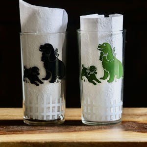 May include: Two clear drinking glasses with white paper napkins inside. Each glass features a dog design, one in black and the other in green. The glasses are on a wooden surface against a dark background.