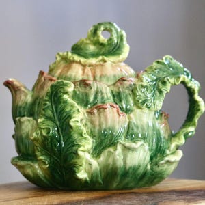 May include: A ceramic teapot with a unique artichoke design. The teapot is primarily green with accents of tan and brown. The handle and spout are leaf-shaped, and the lid has a small hole. The teapot sits on a wooden surface.