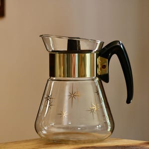 May include: A clear glass coffee carafe featuring a gold-toned metallic band around the upper neck and a black plastic handle. The glass body is decorated with a repeating pattern of gold-colored starburst designs scattered across the exterior surface.
