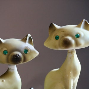 May include: Two vintage cat figurines with cream-colored bodies, dark brown accents, and emerald green eyes. The cat figurines have a mid-century modern aesthetic and are posed upright, with a slender neck and pointed ears.