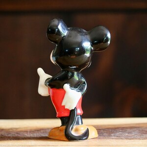 May include: A ceramic figurine of Mickey Mouse, viewed from the back. The figure has a glossy black head and ears, white gloves, red shorts, and yellow shoes. The figurine is standing on a wooden surface against a dark brown background.