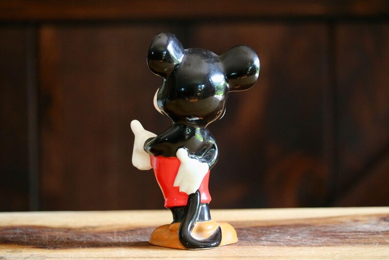 May include: A ceramic figurine of Mickey Mouse, viewed from the back. The figure has a glossy black head and ears, white gloves, red shorts, and yellow shoes. The figurine is standing on a wooden surface against a dark brown background.