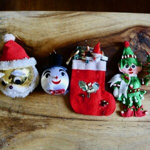 May include: A collection of vintage Christmas ornaments, including a Santa Claus head, a snowman, a red stocking, and an elf holding a small tree. The ornaments are arranged on a wooden surface.
