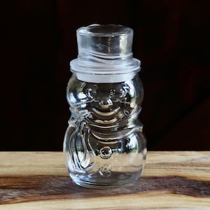 May include: A clear glass snowman-shaped jar with a lid. The jar has a smiling face and a top hat. The jar is sitting on a wooden cutting board against a dark background. The jar is likely for storing candy or small items.